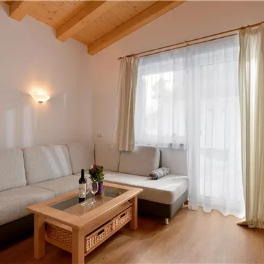 Ein gemütlicher Wohnraum mit einem grauen Sofa und einem Holztisch. Große Fenster lassen viel Licht herein und ein Fernseher ist an der Wand montiert.