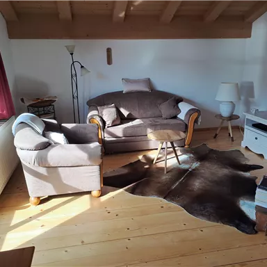 Ein gemütliches Wohnzimmer mit einem Sofa und einem Sessel. Der Raum hat Holzböden und wird von natürlichem Licht erhellt.