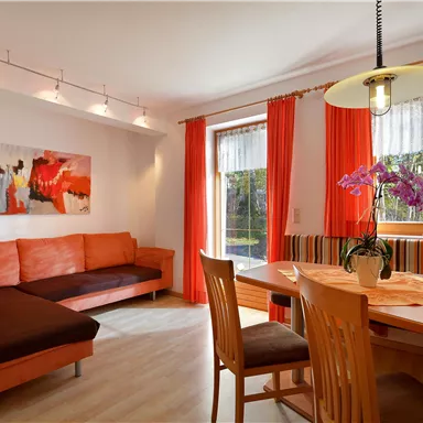 Ein modernes Wohnzimmer mit einer orangefarbenen Couch und einem Essbereich. Große Fenster lassen viel Tageslicht herein und ein paar Pflanzen verschönern den Raum.