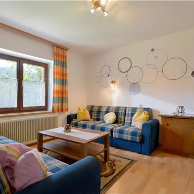 Ein gemütliches Wohnzimmer mit blauen Sofas und bunten Kissen. Große Fenster lassen viel Licht herein und ein Holzschrank ergänzt die Einrichtung.