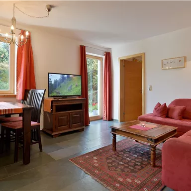 Ein gemütliches Wohnzimmer mit einem roten Sofa und einem Holztisch. Große Fenster lassen viel Licht herein und bieten einen Blick ins Freie.