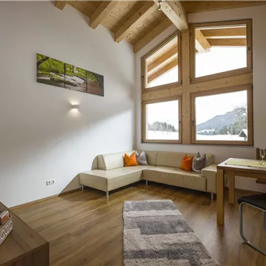 Eine helle und moderne Wohnungecke mit großen Fenstern und viel natürlichem Licht. Es gibt eine gemütliche Sitzecke und einen Esstisch.