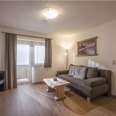 A cozy living room with a gray sofa and a low table. The room has large windows that let in plenty of light and a warm, inviting atmosphere.