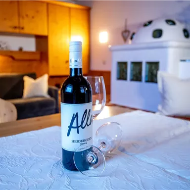 A bottle of red wine with a glass on a table. In the background, a cozy living atmosphere can be seen.