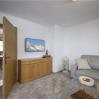 A bright living room with a comfortable sofa and a TV on the wall. In the background, additional furniture and a painting can be seen.