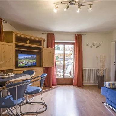 Ein helles Wohnzimmer mit einer blauen Couch und einem Esstisch. Große Fenster bieten Blick auf einen Außenbereich.