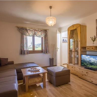 Ein gemütliches Wohnzimmer mit einem großen Sofa und Holzregalen. Ein Fernseher zeigt eine Landschaft, während das Licht von einem Kronleuchter sanft strahlt.