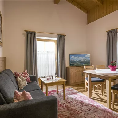 Ein gemütliches Wohnzimmer mit einem grauen Sofa und einem Holztisch. Große Fenster sorgen für viel natürliches Licht.