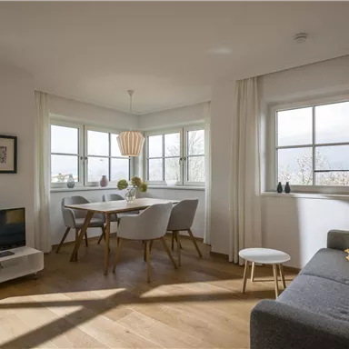 A cozy living room with a gray sofa and a small table. The dining area is bright and inviting, with a view through large windows.