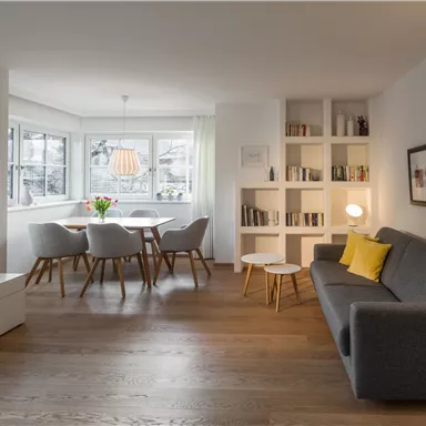 Cozy living room with a gray sofa and modern lighting. A dining table with chairs and shelves in the background create an inviting atmosphere.