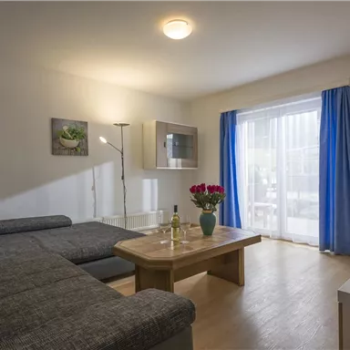 Ein gemütliches Wohnzimmer mit einem grauen Sofa und einem Holztisch. Große Fenster mit blauen Vorhängen sorgen für viel Licht.