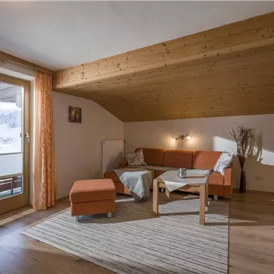 A cozy living room with an orange sofa and a small table. Large windows offer a view of the mountains and daylight comes in.