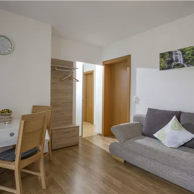 Ein helles Wohnzimmer mit einem grauen Sofa und einem Tisch mit Stühlen. An der Wand hängt ein Bild einer Landschaft, und es gibt eine Uhr sowie einen Kleiderständer.