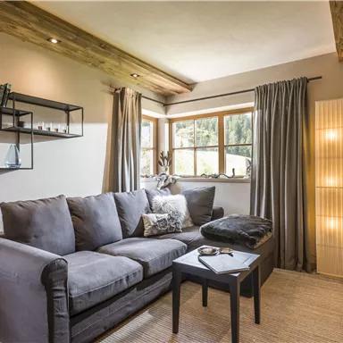 Ein gemütliches Wohnzimmer mit einem grauen Sofa und einem kleinen Tisch. Große Fenster lassen viel Tageslicht hereinfalle, und moderne Dekoration verleiht dem Raum Stil.