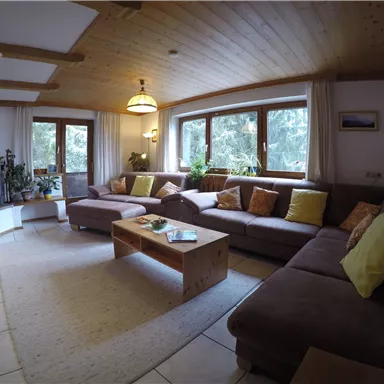 Ein gemütliches Wohnzimmer mit einer U-förmigen Couch und bunten Kissen. Große Fenster lassen viel Licht herein und bieten einen Blick auf die Natur.