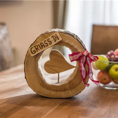 Eine hölzerne Dekoration mit dem Schriftzug "GRIASS DI" und einem Herz in der Mitte steht auf einem Tisch. Im Hintergrund sieht man eine Schüssel mit verschiedenen Früchten.