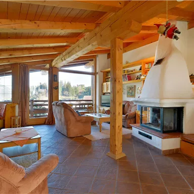 A cozy living space with wooden beams and large windows. The room features comfortable furniture and a fireplace.