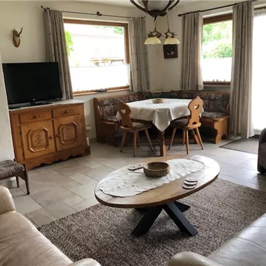 Ein gemütliches Wohnzimmer mit einem großen Tisch und Holzmöbeln. Die Fenster lassen viel Tageslicht herein und der Raum wirkt einladend.