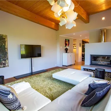 A modern living room with a white sofa, a television, and a fireplace. The room has large windows and a warm wooden ceiling.