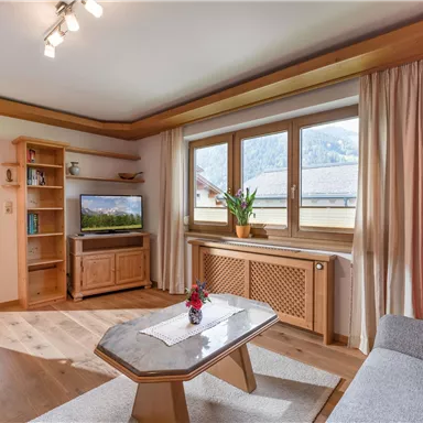 Ein modernes Wohnzimmer mit einem gemütlichen Sofa und einem Fernseher. Große Fenster bieten Blick auf die Berge und viel Tageslicht.