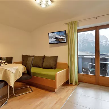 A modern living room with a dining table and a cozy couch. The large window front offers a view of the mountains.