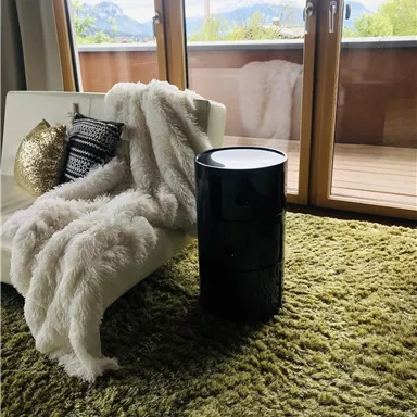 A modern living room with a white sofa, a cozy blanket, and a black side table. Large windows offer a view of nature in the background.