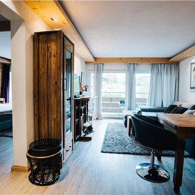 A cozy living room with a dining table, a couch, and large windows. The room is bright and modernly furnished, with wooden and fabric elements.