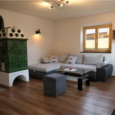 Ein gemütlicher Wohnraum mit einer modernen Couch, einem stilvollen Tisch und einem traditionellen Ofen. Der Raum hat große Fenster, die viel Tageslicht hereinlassen.