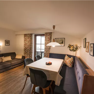 A cozy living room with a large dining table and a comfortable seating area. Windows with floral curtains create an inviting atmosphere.