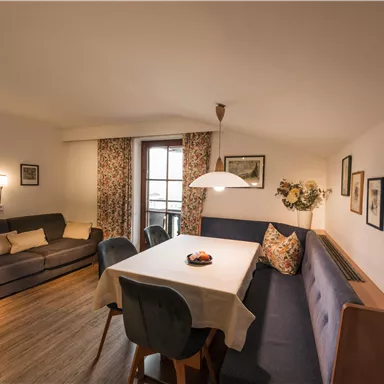 A cozy living room with a large dining table and a comfortable seating area. Windows with floral curtains create an inviting atmosphere.