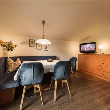 A cozy living space with a seating area and a dining table. The room is bright and cheerful, featuring a television and a dresser.