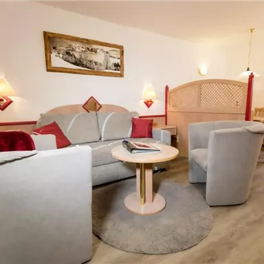 Ein gemütlicher Wohnbereich mit einem Sofa, zwei Sesseln und einem runden Tisch. Die Wände sind in warmen Farben gehalten und es gibt sanfte Beleuchtung.