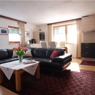 Ein gemütliches Wohnzimmer mit einem schwarzen Sofa und einem Tisch. Im Hintergrund sind eine moderne Küche und große Fenster, die Licht hereinlassen.