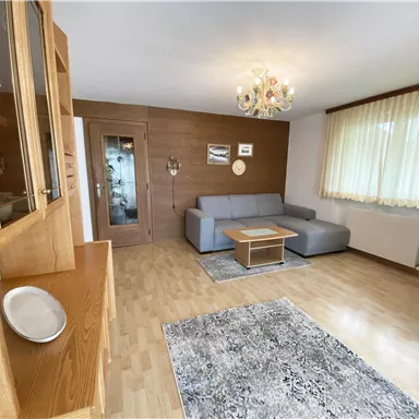 Ein helles Wohnzimmer mit einem grauen Sofa und einem Holztisch. Große Fenster sorgen für viel Tageslicht und eine gemütliche Atmosphäre.