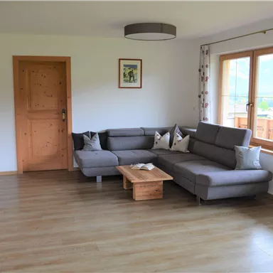 Ein helles Wohnzimmer mit einem grauen Sofa und einem Couchtisch aus Holz. Große Fenster bieten einen Blick nach draußen und lassen viel Licht herein.