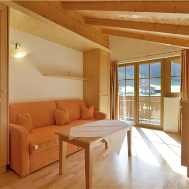 Ein gemütliches Wohnzimmer mit einer orangenen Couch und einem Holztisch. Große Fenster sorgen für viel Tageslicht und Ausblick nach draußen.