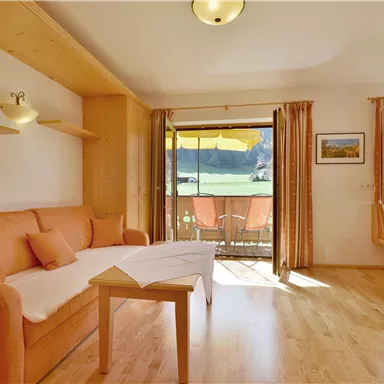 Ein gemütliches Wohnzimmer mit einem Sofa und Holzmöbeln. Durch die Fenster, die zur Terrasse führen, fällt viel Tageslicht herein.