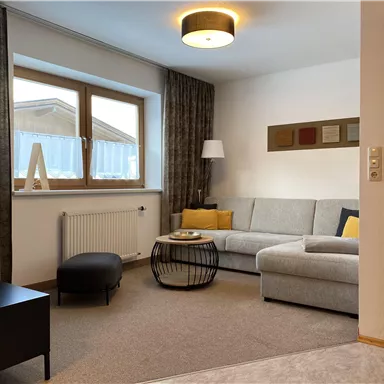 Ein gemütliches Wohnzimmer mit einem grauen Sofa und einem runden Couchtisch. Große Fenster lassen viel Licht herein und die Dekoration ist modern und stilvoll.