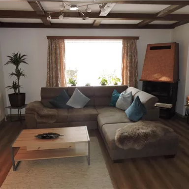 Ein gemütliches Wohnzimmer mit einem großen, braunen Sofa und blau-grünen Kissen. Es gibt einen Fenster mit Vorhängen, eine Pflanze in der Ecke und einen Holzofen.