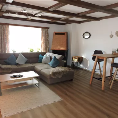 Ein gemütliches Wohnzimmer mit einer großen Couch, einem Tisch und Stühlen. Es gibt ein Fenster mit Vorhängen und eine warme Holzdecke.