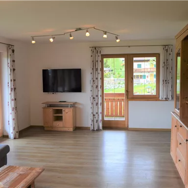 Ein modernes Wohnzimmer mit einem Fernseher und großen Fenstern. Die Möbel sind aus hellem Holz und die Atmosphäre ist hell und einladend.