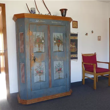 A traditional cabinet with colorful floral patterns stands in a bright entrance area. Next to the cabinet is a red wooden chair.