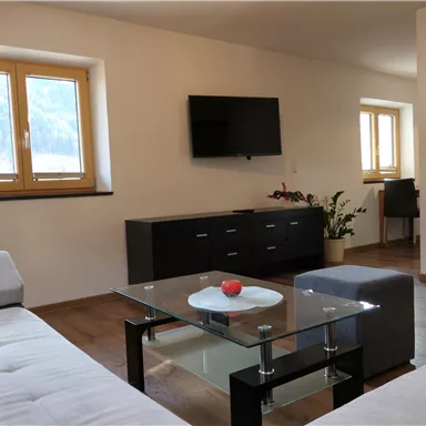 Ein modernes Wohnzimmer mit einem weißen Sofa, einem Glastisch und einem Fernseher an der Wand. Große Fenster lassen viel natürliches Licht herein.