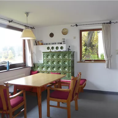 A cozy dining area with a wooden table and chairs in a bright room. Large windows let in plenty of daylight and offer a view outside.