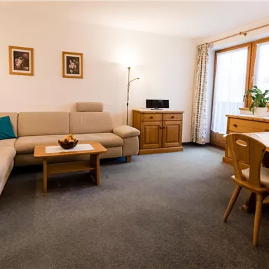 Ein helles Wohnzimmer mit einem beigen Sofa und einem Holztisch. Der Raum ist gemütlich und hat große Fenster mit Blick nach draußen.