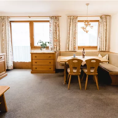 Ein gemütliches Wohnzimmer mit Holzmöbeln und einem Essbereich. Große Fenster lassen viel Tageslicht herein.