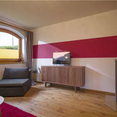 A modern living room with a gray sofa and a wood stove. A television stands on a wooden cabinet and a red wall provides an accent.
