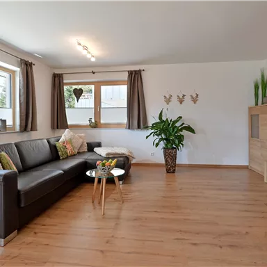 Ein modernes Wohnzimmer mit einem schwarzen Sofa und einem Holztisch. Große Fenster lassen viel Licht herein und es gibt Pflanzen zur Dekoration.