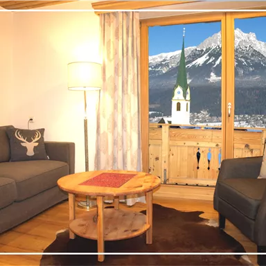 Ein gemütliches Wohnzimmer mit einem grauen Sofa und einem Holz-Couchtisch. Durch das Fenster sieht man eine beeindruckende Berglandschaft und einen Kirchturm.