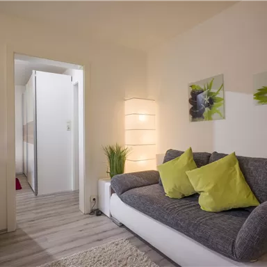 A modern living room with a gray sofa and light green cushions. The room is brightly lit and has a friendly atmosphere.